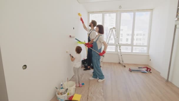 家庭时间与小女儿在一起 年轻的父亲和母亲展示他们的孩子如何画墙与滚筒 油漆颜色为浅蓝色 家里的房间准备装修 — 图库视频影像