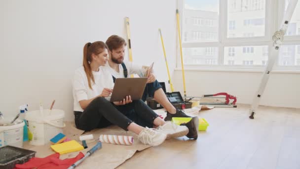 满意的年轻夫妇看着笔记本电脑计划新的内部 坐在地板上白墙背景的空房间里 一对快乐的年轻夫妇在他们的新房子里计划着如何修理笔记本电脑 修理的想法 — 图库视频影像