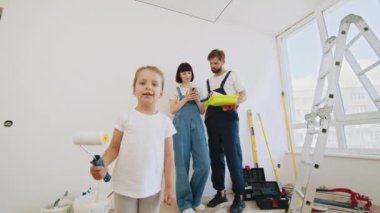 Kameraya gülümseyen, elinde boya fırçasıyla odada duran sevimli, mutlu küçük kızın portresini yakından çek. Annemle babam arka plana renkli duvar boyuyor. Ev tamiri. İç tasarım.