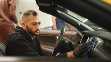 Cleaning service, car detailing concept. Young man, auto service worker, washing a car interior and dashboard with microfiber cloth, in a professional car wash. Service worker, washing a car interior