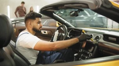 Close up portrait of professional car wash service worker in overalls, white t-shirt and protective gloves cleaning car interior and control panel with special chemical anti dust liquid and soft brush