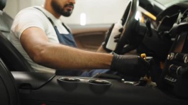 Bearded young male car detailing worker in overalls, sitting inside of modern vehicle, and cleaning car dashboard, putting plastic trim care milk on the soft brush