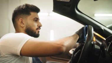 Handsome bearded young man in uniform and protective gloves, cleaning car interior and steering wheel using microfiber cloth, smiling at camera. Car detailing and valeting concept.