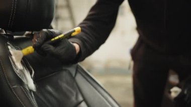 Car detailing and car wash concept. Close up hands of man, car wash worker, wearing black clothes and protective rubber gloves, cleaning car front seat, spraying the foam on the soft brush
