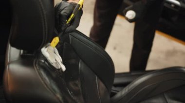 Close up cropped image of washing car interior and seat by a soft brush with foam. Young male worker in black protective gloves cleaning car seat with foam and brush. Washing car interior