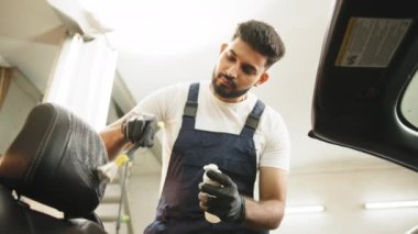 Close up cropped image of washing car interior and seat by a soft brush with foam. Young male worker in overalls and black protective gloves cleaning car seat with foam and brush. Cleaning car
