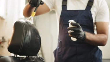 Close up cropped image of washing car interior and seat by a soft brush with foam. Young male worker in overalls and black protective gloves cleaning car seat with foam and brush. Washing car interior