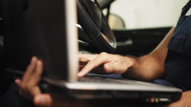 Cropped view of bearded male mechanic sitting inside car using laptop, recording automobile engine checks collect detailed information during his work on car workshop. Male mechanic inside car.