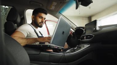 Genç sakallı adam araba teknisyeni kırmızı arabanın içinde oturmuş motor problemini tamir ediyor, sistem kontrolü sırasında, garajda bakım ve onarım için dizüstü bilgisayar kullanıyor..