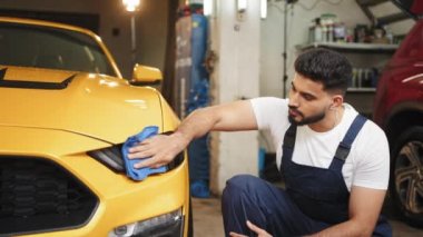 Bearded car wash worker man, wearing protective clothes uniform overalls, cleaning modern yellow luxury car with microfiber cloth. Car detailing or vehicle concept. Bearded car wash worker man