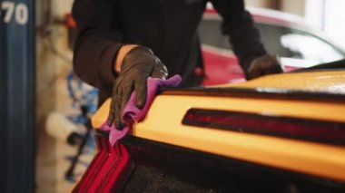 Cropped view of hand of male car wash worker holds the microfiber and polishes yellow sport luxury car under tail lights of vehicle. Car detailing and cleaning concept. Hand of male car wash worker.