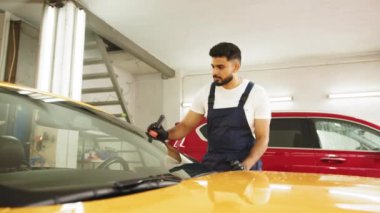 Mavi tulumlu, koruyucu eldivenli, profesyonel bir araba yıkama servisine odaklan. Lüks sarı arabanın ön camına yağmur önleyici sür..