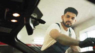 Otomobil servisinde yağmur önleyici kaplama. Mavi tulumlu ve koruyucu eldivenli bir erkek işçi profesyonel bir araba yıkama servisinde, lüks sarı arabanın ön camına yağmur önleyici sürüyor..