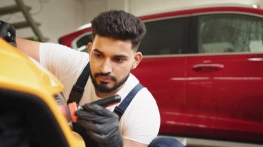 Tulumlu ve koruyucu eldivenli çekici genç sakallı adam, araba temizleme servisinde sarı spor araba cam farları için yağmur önleyici kompozisyon uyguluyor..