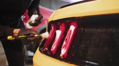Close up of young male worker in black protective gloves cleaning tail light in foam with special brush. Sport yellow luxury car wash and detailing. Washing machine at the station.