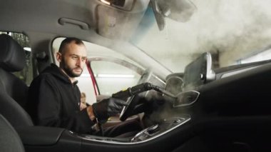 Cleaning of car salon with steam cleaner. Side view of professional male confident worker cleaning car interior, steering wheel and control panel using hot steam cleaner. Cleaning interior of the car