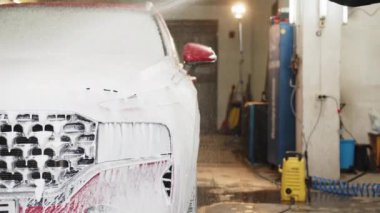 El yapımı araba yıkama konsepti. Genç işçi, benzin istasyonunda modern kırmızı araba kaputunu köpükle ve basınçlı suyla temizliyor. El yıkaması, basınçlı su ve oto yıkama.