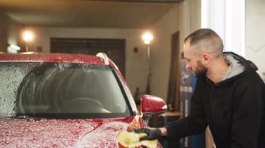 Young bearded car service male worker doing cleaning the hood of red crossover car with the help of shampoo and yellow sponge in car washing company. Car washing service.