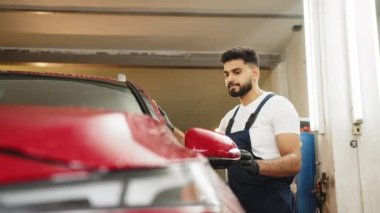 Araba yıkama ve detay verme konsepti. Genç konsantre sakallı erkek işçi süngeri elinde tutuyor ve kırmızı araba kaputunu sabunla temizliyor..