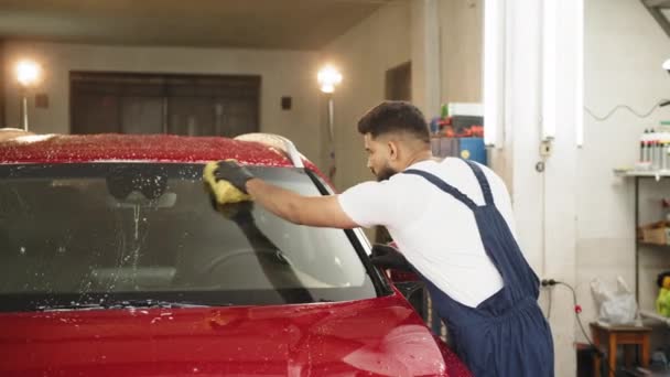 年轻的胡子男子 洗车工人 穿着T恤和工作服 借助特殊的泡沫和海绵清洗汽车挡风玻璃 用手洗汽车 用海绵和泡沫泡 — 图库视频影像