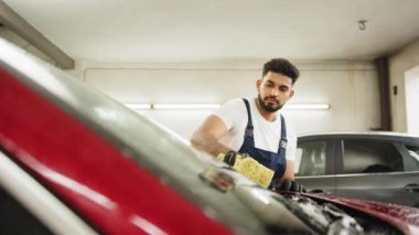Genç sakallı bir adam, araba yıkama işçisi, tişört giyiyor ve özel köpük ve süngerle arabanın ön camını temizliyor. Arabayı elle yıkamak, sünger ve köpük köpükleri.