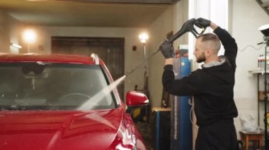 Araba yıkama servisinde yüksek basınçlı su altında kırmızı araba yıkayan genç sakallı adam portresi. Sabunu suyla temizlemek, araba yıkamak..