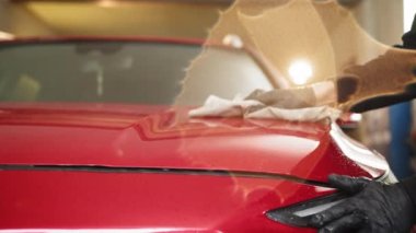 Car detailing and cleaning concept. Good-looking young male car wash worker, wearing special clothes and gloves, wipes and polishes the car hood with microfiber cloth. Car wash service.