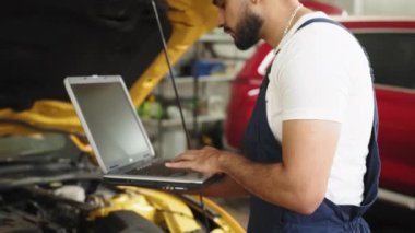Bearded male mechanic using laptop, recording automobile engine checks collect detailed information during his work on car workshop. Service maintenance during engine repair. Service maintenance.