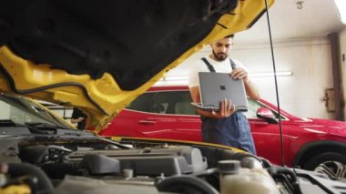 Close up of young bearded engineer mechanic under car hood checking engine with laptop using electronic diagnostic equipment to tune sport yellow vehicle. Service maintenance during engine repair
