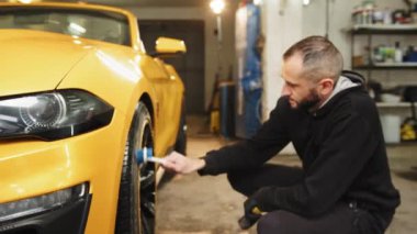 Close up of handsome young male employee worker cleaning the wheel tire of modern yellow sport car by scrubbing brush. Manual wash at car wash maintenance service.