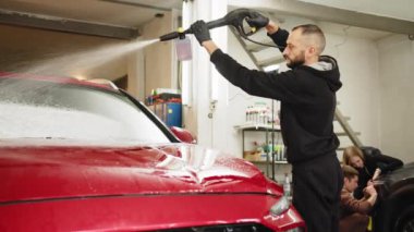Bearded young man car wash worker spraying cleaning foam to a modern red car holding a high pressure washer. Modern car and foam washing. Young man car wash worker spraying cleaning