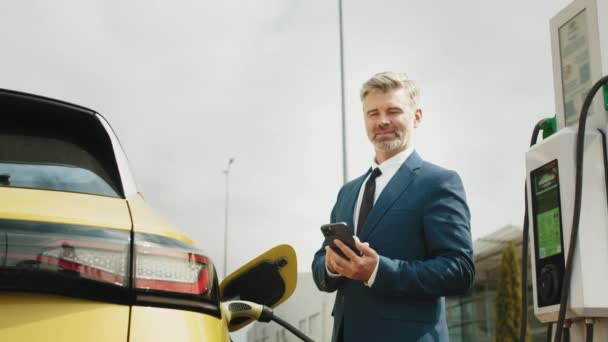 Geschäftsmann Hält Smartphone Während Auto Ladestation Für Elektrofahrzeuge Auflädt Nahaufnahme — Stockvideo
