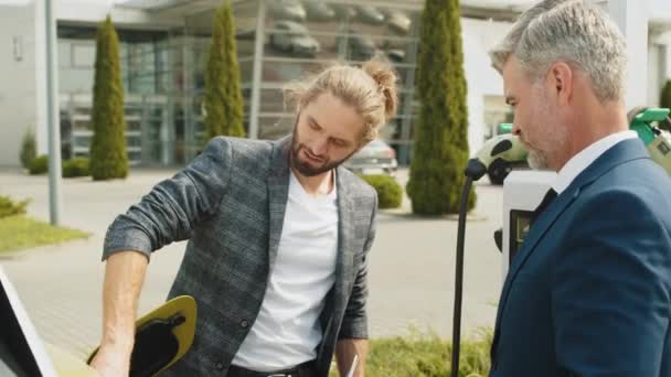 两名男子在汽车充电站附近 年轻自信的推销员展示了在户外为顾客充电的过程 灰发男人在买电动车 环境友好型现代绿色交通 — 图库视频影像