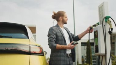 Halka açık bir şarj istasyonunda beyaz bir genç adam elektrikli araba şarj ediyor. Ekoloji alternatif enerjisini geleceğin sürdürülebilir enerjisinden koru. Elektronik araç EV modern otomobili şarj ediyor