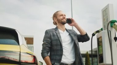 Genç yakışıklı adam, istasyonda elektro arabası şarj olurken telefonu kullanıyor. Elektrikli araba şarj konsepti. Şarj istasyonunda şarj kablosu olan gülümseyen adam.