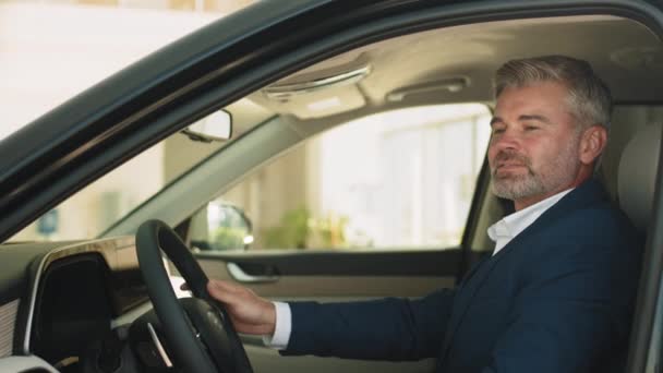 成功したビジネスマンは運転席に座って購入を楽しむ 幸せなひげ灰色の髪の男はついに待望の車を取得し 正式なスーツを着て ディーラー店 販売コンセプト — ストック動画