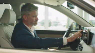 Erkek, otomobil kabininde oturuyor ve yeni arabayı test ediyor. Medya ekranını kullanıyor, dokunuyor, parmağıyla işaret ediyor. Resmi olarak başarılı bir iş adamı sürücü koltuğunda oturuyor. Gözlere odaklan.