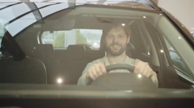 Yeni arabasında oturan mutlu, gülümseyen genç bir alıcı bayi otoparkının dışında izole edilmiş geziye hazır bekliyor. Araba galerisindeki arabaya bakan bir adam. Otomatik satın alma yetkileri.