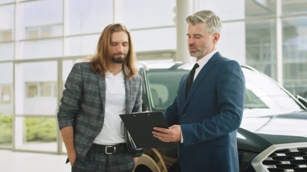 快乐的专业推销员和客户为汽车的销售或租赁签署文件 成功的中年商人在一家汽车展示室里把汽车卖给男性买主 — 图库视频影像