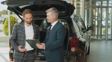 Kendine güveni tam, gri saçlı satıcı tüm araba özelliklerini yakışıklı sahibine açıklıyor. Gri saçlı adam galerideki müşteriye yepyeni bir araba gösteriyor.