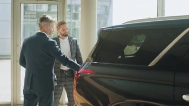 Kendine güveni tam, gri saçlı satıcı tüm araba özelliklerini yakışıklı sahibine açıklıyor. Müşteri aracın bagajının içine bakıyor. Otomatik çapraz bagaj.