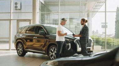 Beyaz tişörtlü yakışıklı, olgun bir adam araba alıyor, modern bir araba galerisinde banka kartı olan bir terminal kullanıyor. Adam almak istediği otomobili seçer. Satış konsepti.