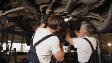 Üniformalı iki tamirci baba ve oğlu kaldırılmış araçla otomobil servisinde çalışıyor. Araba asansörde. Profesyonel oto tamircisi bir arabanın şasisi üzerinde çalışıyor.