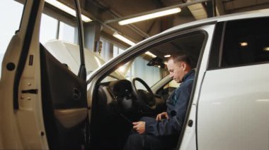 Bir tamirci atölyede dizüstü bilgisayarla araba taraması yapıyor. Otomobil servisi, bakım konsepti. Tamirci arabayı kontrol ediyor, benzin istasyonunda dizüstü bilgisayarla tarama yapıyor. Bilgisayar kullanan bir adam.