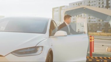 Dışarıdaki şarj istasyonunda beyaz lüks elektro arabasına elektrik kablosu takan şık bir işadamının arka görüntüsü. Ulaşım, ekoloji ve modern yaşam tarzları kavramı. Elektrikli araba yükleniyor..