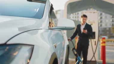 Gülümseyen Asyalı işadamı elektrik kablosunu şarj ederken akıllı telefon ekranına bakıyor. İnsan EV istasyonunda duruyor ve modern ve yenilikçi teknolojilerle yaşam tarzının tadını çıkarıyor.