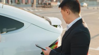 Siyah takım elbiseli Asyalı bir adamın beyaz lüks arabayı şarj edip modern akıllı telefon kullanmasına yakın çekim. Elektrikli araç teknolojik şarj konsepti. Arabaları şarj eden ve akıllı telefonları kullanan adama yaklaş..