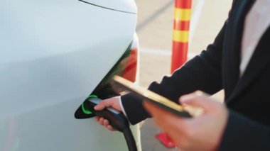 Erkek eliyle elektrik kablosunu açık hava elektrik santralinde beyaz elektrikli arabaya yerleştiriyor. Arka plan bulanık. Bilinçli sahibi olan çevre dostu bir nakliye aracı. Adam arabayı şarj ediyor ve akıllı telefon kullanıyor..