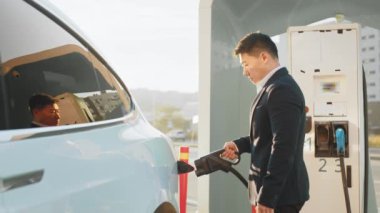 Adam arabayı şarj ediyor. Modern elektro arabanın yanında duran şık giyimli kendine güvenen Asyalı adamın yan görünüşü. Yakışıklı erkek hava kirliliğini önlemek için çevre dostu araç kullanıyor