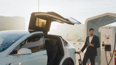 İstasyondaki Hintli adam elektrikli arabayı şarj ediyor. Takım elbiseli Arap işadamı bir elinde hazır kahveyle dışarıda dikilirken beyaz elektrikli arabaya elektrik kablosu sokuyor. Sıfır emisyon aracı